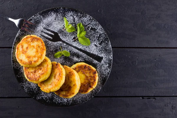 Cottage cheese pancakes on a dark background. Syrniki with fresh mint. Pancakes with cottage cheese on a black plate sprinkled with powdered sugar. Healthy food