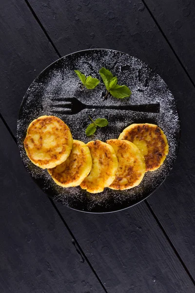 Cottage cheese pancakes on a dark background. Syrniki with fresh mint. Pancakes with cottage cheese on a black plate sprinkled with powdered sugar. Healthy food