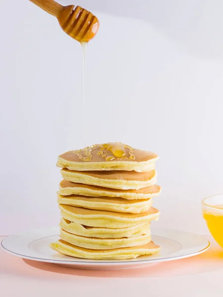 Pancakes on a white pink background. Hot pancakes with honey on a white plate. Delicious dish for breakfast. Honey flows down from a wooden stick on a dish. Healthy food