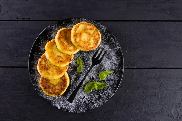 Cottage cheese pancakes on a dark background. Syrniki with fresh mint. Pancakes with cottage cheese on a black plate sprinkled with powdered sugar. Homemade food