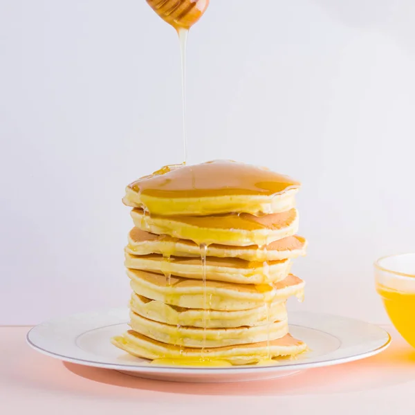 Pancakes on a white pink background. Hot pancakes with honey on a white plate. Delicious dish for breakfast. Honey flows down from a wooden stick on a dish. Homemade food