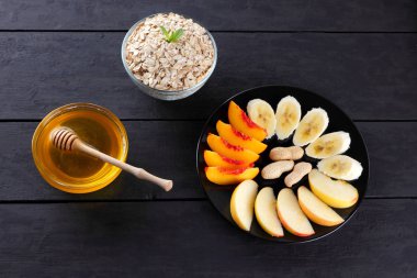 Yulaf ezmesi, bal ve kahvaltı için nane ile dilimlenmiş meyve. Yulaf gevreği, muz dilimleri, dilimlenmiş elma ve şeftali. Yulaf ezmesi, çeşitli meyve ve siyah ahşap kurullarında tatlım. Sağlıklı gıda