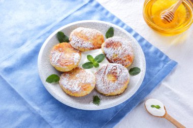 Süzme peynir pudra şekeri serpilir ve taze nane krep. Cheesecakes beyaz tabakta. Tahta kaşık ekşi krema. Beyaz mavi zemin üzerine bal ve süt ürünleri. Sağlıklı gıda