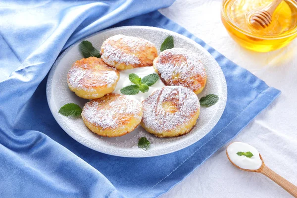 Cottage cheese pancakes sprinkled powdered sugar and fresh mint. Cheesecakes on white plate. Sour cream in wooden spoon. Honey and dairy products on white blue background. Healthy food