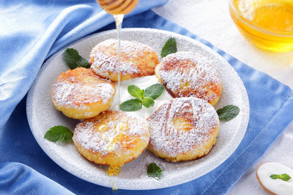 Cottage cheese pancakes sprinkled powdered sugar and fresh mint. Cheesecakes on white plate. Sour cream in wooden spoon. Honey and dairy products on white blue background. Healthy food