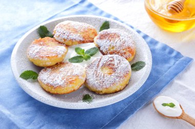 Süzme peynir pudra şekeri serpilir ve taze nane krep. Cheesecakes beyaz tabakta. Tahta kaşık ekşi krema. Beyaz mavi zemin üzerine bal ve süt ürünleri. Sağlıklı gıda