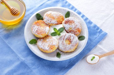 Süzme peynir pudra şekeri serpilir ve taze nane krep. Cheesecakes beyaz tabakta. Tahta kaşık ekşi krema. Beyaz mavi zemin üzerine bal ve süt ürünleri. Sağlıklı gıda