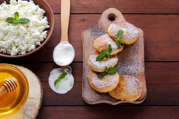 Cottage cheese pancakes sprinkled with powdered sugar. Cheesecakes on wooden board. Cottage cheese in clay bowl and sour cream in wooden spoon. Honey and dairy products. Copy space