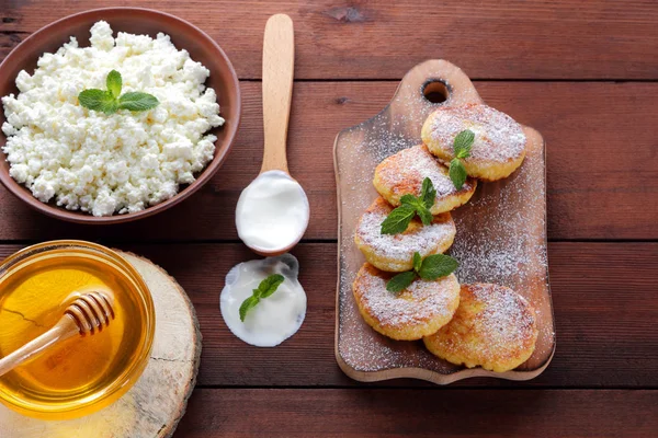 Cottage cheese pancakes sprinkled with powdered sugar. Cheesecakes on wooden board. Cottage cheese in clay bowl and sour cream in wooden spoon. Honey and dairy products. Copy space