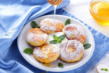 Süzme peynir pudra şekeri serpilir ve taze nane krep. Cheesecakes beyaz tabakta. Tahta kaşık ekşi krema. Beyaz mavi zemin üzerine bal ve süt ürünleri. Sağlıklı gıda
