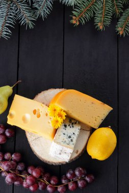 Various types of cheese and fruits on dark boards and Christmas tree branches. Swiss cheese, dorblu, Dutch cheese for Christmas. Dairy products, lemon, grapes, pears. Top view clipart