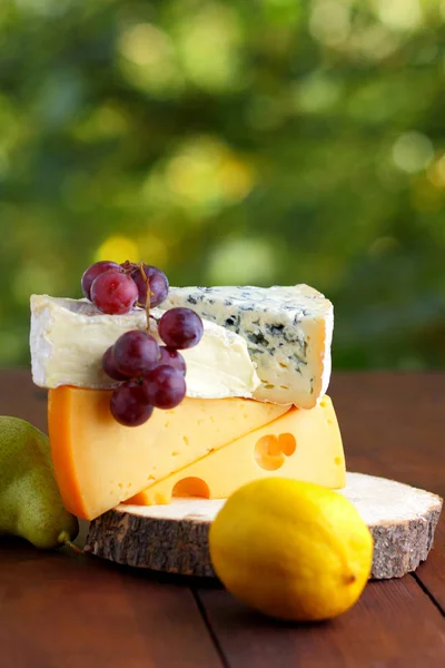 Camembert, brie, hard cheese and grapes on a wooden stand. Pieces of different cheeses, pear and lemon on a blurred background. Dorblu and soft cheese on wooden boards. Cheese and fruit on bokeh