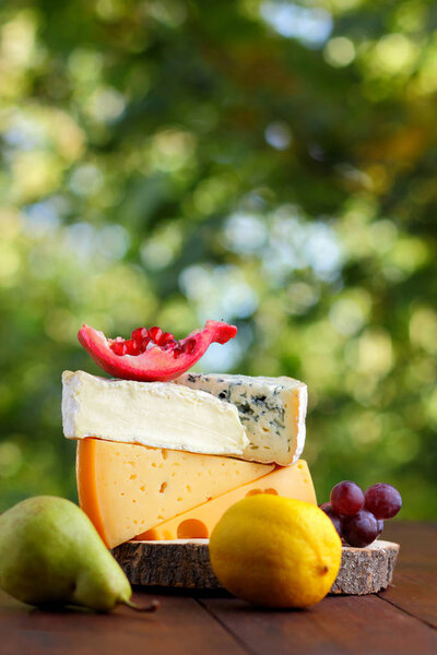 Camembert, brie, hard cheese and grapes on a wooden stand. Pieces of different cheeses, pear and lemon on blurred background. Dorblu, soft cheese and pomegranate on boards. Cheese and fruit