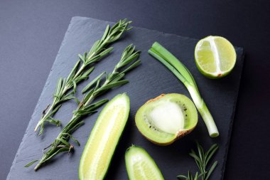 Yeşil meyve ve sebze karanlık kayrak gemide. Doğal yeşil ürünler kavramı. Avokado, kivi, limon ve elma koyu arka plan üzerinde. Rosemary, dereotu ve Frenk soğanı taş tahta üzerinde