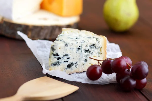 Various types of cheese, pear and grapes on wooden board. Slices of various cheeses and grapes on blurred background. Dorblu, camembert and hard yellow cheese on parchment paper