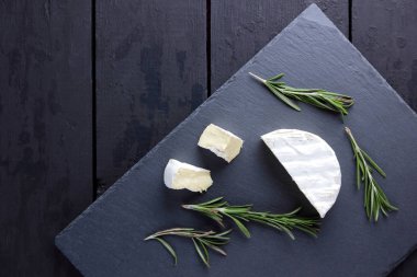 Camembert ve biberiye siyah taş gemide. Siyah arka plan üzerine beyaz kalıp ile yumuşak peynir. Kayrak kurulu dilimlenmiş peynir ve biberiye dallarda
