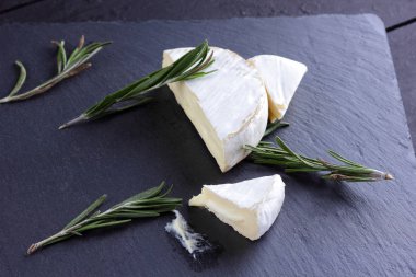 Camembert ve biberiye siyah taş gemide. Siyah arka plan üzerine beyaz kalıp ile yumuşak peynir. Kayrak kurulu dilimlenmiş peynir ve biberiye dallarda