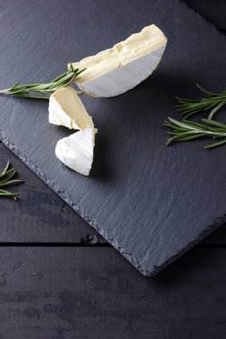 Camembert ve biberiye siyah taş gemide. Siyah arka plan üzerine beyaz kalıp ile yumuşak peynir. Kayrak kurulu dilimlenmiş peynir ve biberiye dallarda