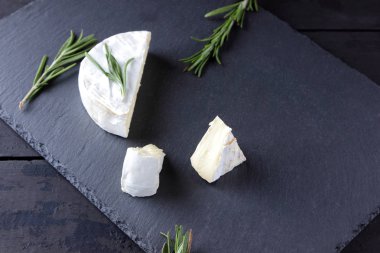 Camembert ve biberiye siyah taş gemide. Siyah arka plan üzerine beyaz kalıp ile yumuşak peynir. Kayrak kurulu dilimlenmiş peynir ve biberiye dallarda