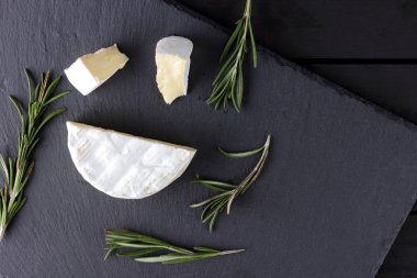 Camembert ve biberiye siyah taş gemide. Siyah arka plan üzerine beyaz kalıp ile yumuşak peynir. Kayrak kurulu dilimlenmiş peynir ve biberiye dallarda