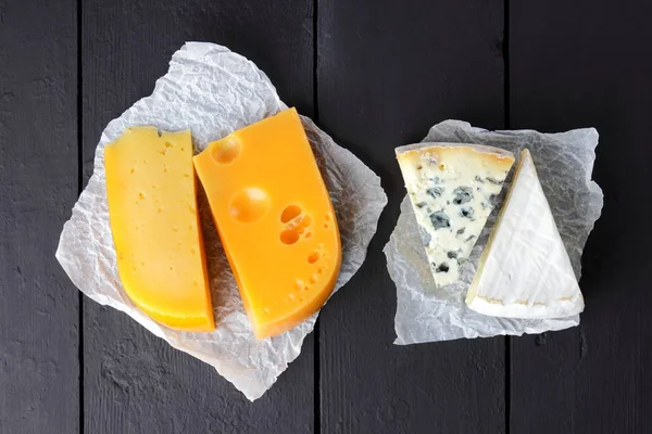 Hard yellow cheese, camembert and blue cheese on black boards. Various types of cheese on parchment paper. Swiss cheese and dorblu on dark background. Top view