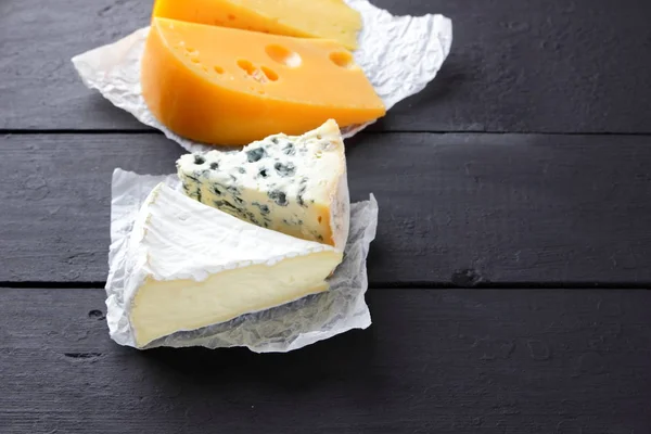 Hard yellow cheese, camembert and blue cheese on black boards. Various types of cheese on parchment paper. Swiss cheese and dorblu on dark background. Top view