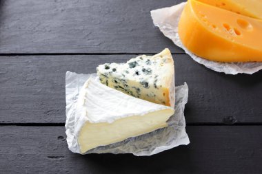 Sert sarı peynir, camembert ve mavi peynir siyah kurullarında. Parşömen kağıt üzerine peynir türleri. İsviçre peyniri ve dorblu koyu arka plan üzerinde. Üstten Görünüm