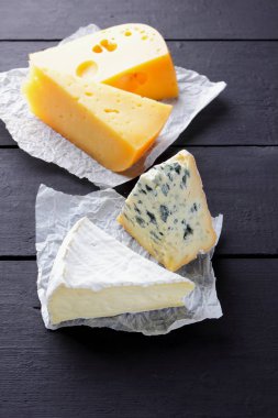 Sert sarı peynir, camembert ve mavi peynir siyah kurullarında. Parşömen kağıt üzerine peynir türleri. İsviçre peyniri ve dorblu koyu arka plan üzerinde. Üstten Görünüm