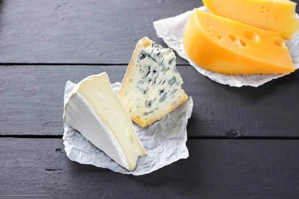 硬黄色奶酪 卡门伯特和蓝色奶酪在黑色的板上 羊皮纸上有各种类型的奶酪 瑞士奶酪和黑暗背景上的背布 顶视图 — 图库照片