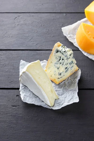 硬黄色奶酪 卡门伯特和蓝色奶酪在黑色的板上 羊皮纸上有各种类型的奶酪 瑞士奶酪和黑暗背景上的背布 顶视图 — 图库照片