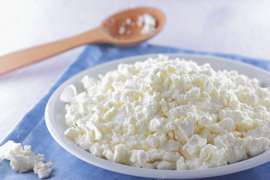 Cottage cheese on blue napkin. Cheese on white plate with wooden spoon. Soft cheese for diet. Healthy dairy product for breakfast