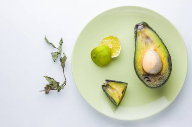 Beyaz arka planda çirkin yiyecek. Yeşil plaka üzerinde çirkin çürük avokado. Bir tabak üzerinde kötü kireç ve kuru nane. Çürük tropikal meyveler. Çürük meyve kavramı. Üstteki görünüm, kopya alanı