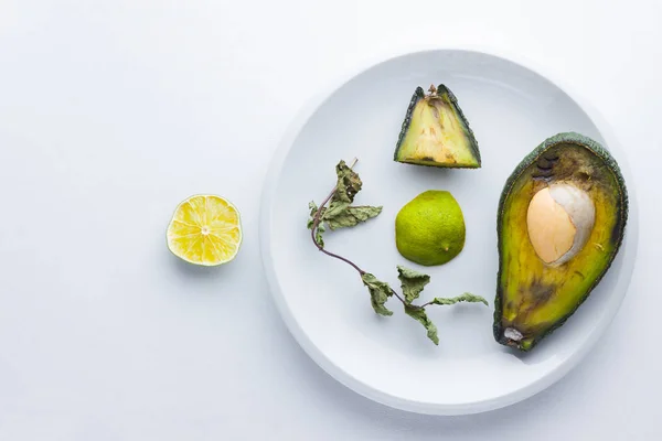 Ugly food on white background. Ugly rotten avocado on white plate. Bad lime and dry mint on a plate. Rotten tropical fruits. Concept of rotten fruit. Top view, copy space