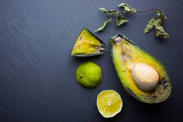 Ugly food on black background. Ugly rotten avocado on slate board. Bad lime and dry mint on black slate board. Rotten tropical fruits. Concept of rotten fruit. Top view, copy space