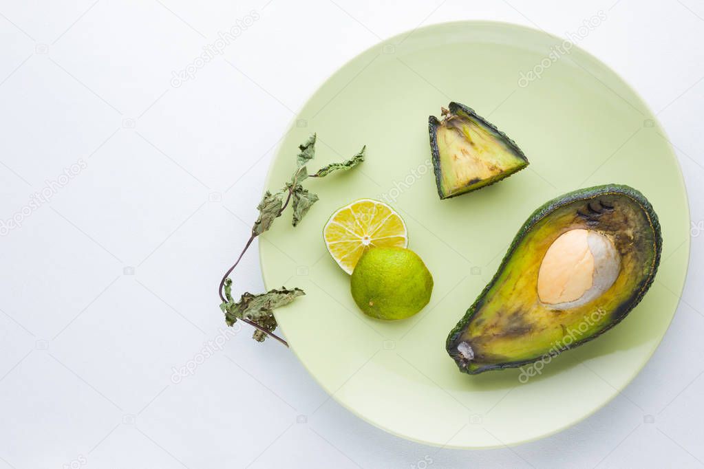 Ugly food on white background. Ugly rotten avocado on green plate. Bad lime and dry mint on a plate. Rotten tropical fruits. Concept of rotten fruit. Top view, copy space