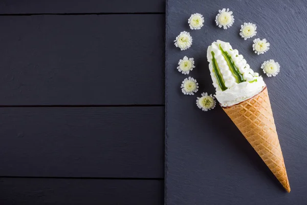 Ice cream cone and white flowers, summer concept. Waffle cone with ice cream on slate board. Dessert with jam on black background. Flat lay