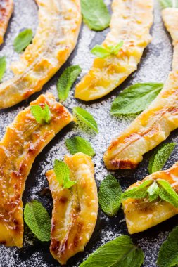 Siyah bir mutfak tahtası ve şist taşı üzerinde pudra şekeri ile ızgara muz yarım. Siyah arka plan masada nane ile Fırında muz tatlısı. Görünümü kapat