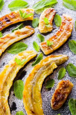 Siyah bir mutfak tahtası ve şist taşı üzerinde pudra şekeri ile ızgara muz yarım. Siyah arka plan masada nane ile Fırında muz tatlısı. Görünümü kapat