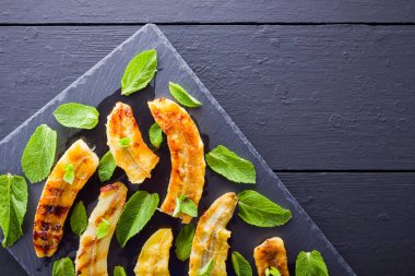 Siyah mutfak tahtası ve şist taşı üzerinde ızgara muz yarım. Siyah arka planda nane ile Fırında muz tatlısı. Asya sağlıklı gıda. Üstte görüntü