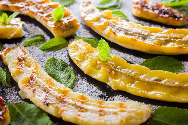 Siyah bir mutfak tahtası ve şist taşı üzerinde pudra şekeri ile ızgara muz yarım. Siyah arka plan masada nane ile Fırında muz tatlısı. Görünümü kapat