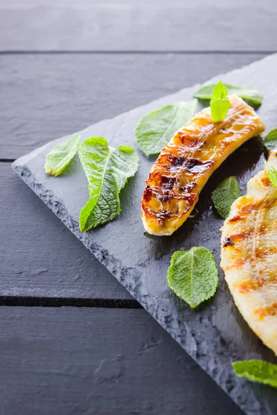 Halves of grilled bananas on a black kitchen board and shale stone. Baked banana dessert with mint on black background. Asian healthy food. Top view