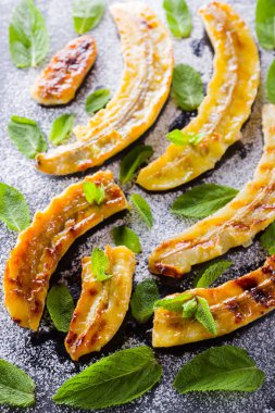 Siyah bir mutfak tahtası ve şist taşı üzerinde pudra şekeri ile ızgara muz yarım. Siyah arka plan masada nane ile Fırında muz tatlısı. Görünümü kapat