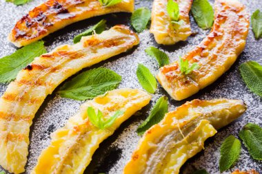 Siyah bir mutfak tahtası ve şist taşı üzerinde pudra şekeri ile ızgara muz yarım. Siyah arka plan masada nane ile Fırında muz tatlısı. Görünümü kapat