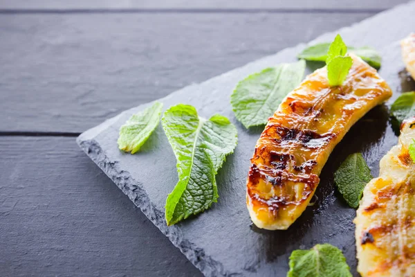 Halves of grilled bananas on a black kitchen board and shale stone. Baked banana dessert with mint on black background. Asian healthy food. Top view