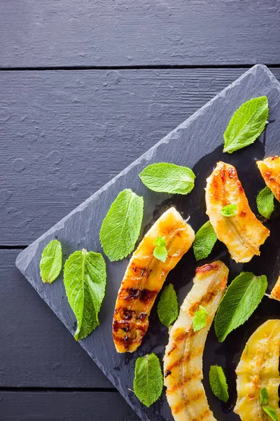 Halves of grilled bananas on a black kitchen board and shale stone. Baked banana dessert with mint on black background. Asian healthy food. Top view