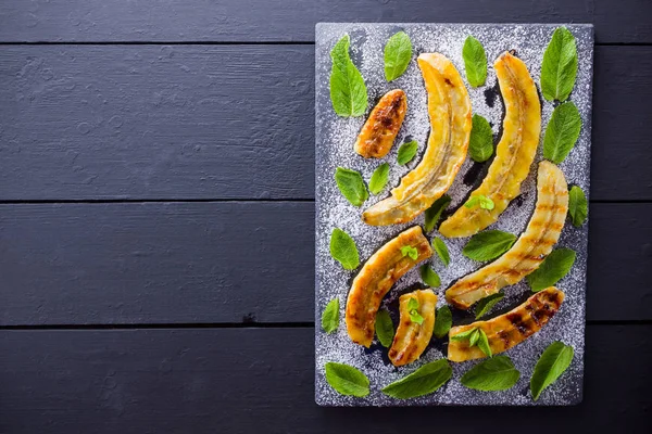 Halves of grilled bananas with powdered sugar on a black kitchen board and shale stone. Baked banana dessert with mint on black background table. Top view