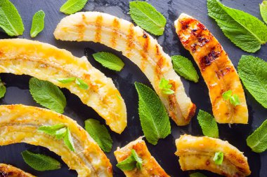 Siyah mutfak tahtası ve şist taşı üzerinde ızgara muz yarım. Siyah arka planda nane ile fırında muz tatlısı. Asya sağlıklı gıda