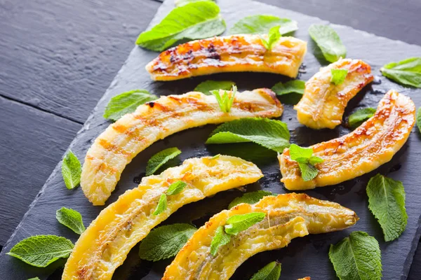 Halves of grilled bananas on a black kitchen board and shale stone. Baked banana dessert with mint on black background. Asian healthy food