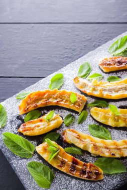 Siyah bir mutfak tahtası ve şist taşı üzerinde pudra şekeri ile ızgara muz yarım. Siyah arka plan masada nane ile Fırında muz tatlısı