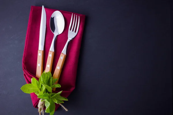 Cutlery set. Knife, spoon, fork on slate stone. utlery with burgundy napkin and twine. Mint on black background. Table setting. Top view